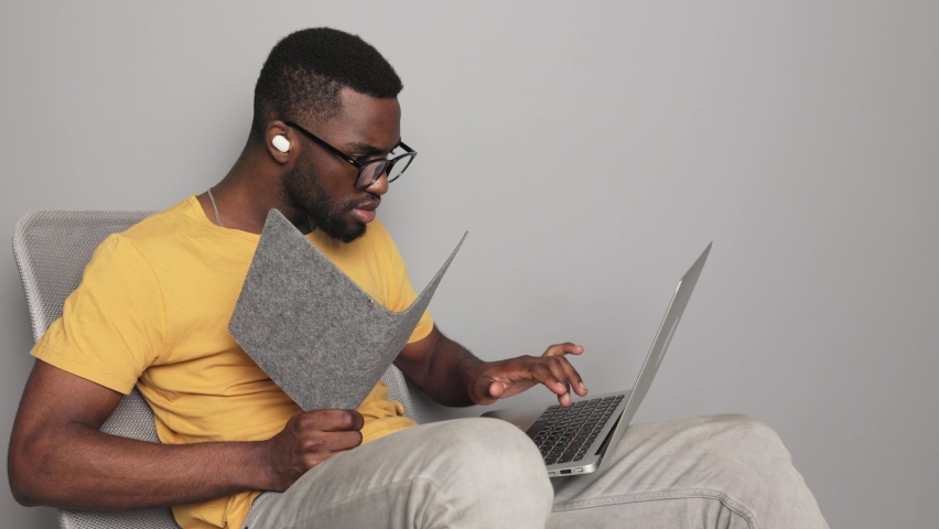 African american man in glasses looks in a notebook student in headphones and works in a laptop checking his knowledge at home online quarantine online training in a yellow t-shirt | Shutterstock HD Video #1094960111