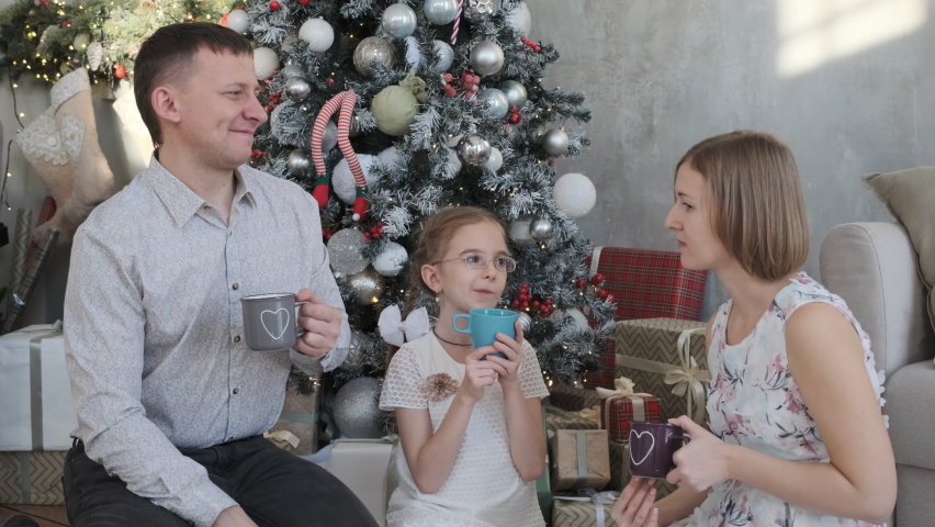 Young family of three drinking hot chocolate or tea while sitting on the floor by the Christmas tree, celebrating winter holidays together | Shutterstock HD Video #1094960335