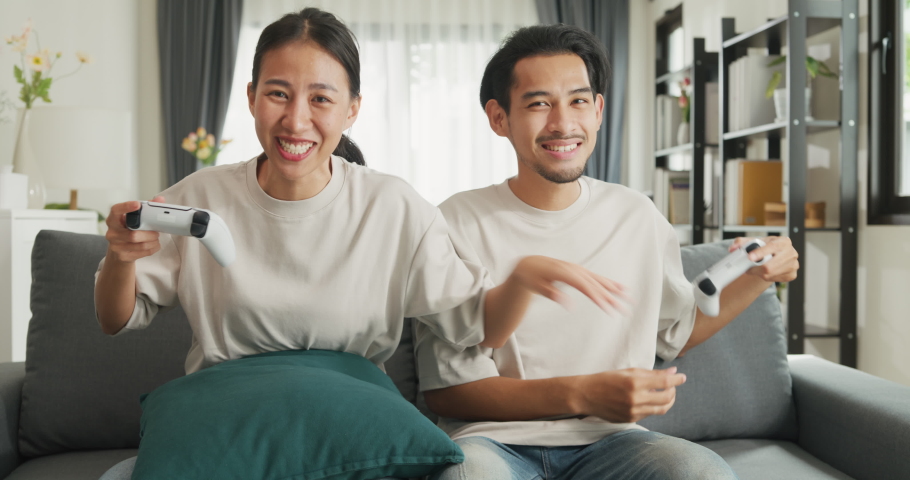 Young Asian couple sit on couch hold joystick play video game spend time together have fun at home on weekend. Happy husband and wife laugh relax with online game, Lifestyle activity concept. | Shutterstock HD Video #1094961435