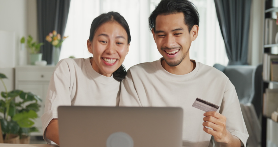 Happy young Asian couple sit on couch with laptop success credit card payment online shopping at home on holiday. Smile young husband and wife excited browsing app on gadgets. Lifestyle concept. | Shutterstock HD Video #1094961437