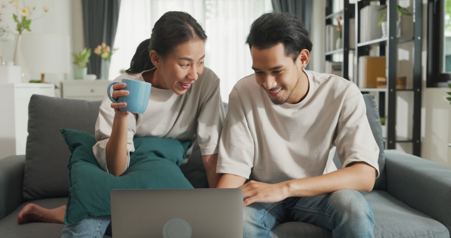 Happy young Asian couple sit on couch fun use laptop computer online shopping at home on holiday. Smiling young husband and wife laugh relax at home browsing application on gadgets, Lifestyle concept. | Shutterstock HD Video #1094961449