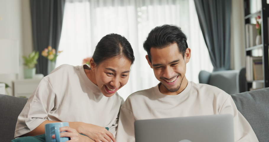 Happy young Asian couple sit on couch use laptop successful payment online shopping at home on holiday. Smile young husband and wife excited at home browsing application on gadgets. Lifestyle concept. | Shutterstock HD Video #1094961457