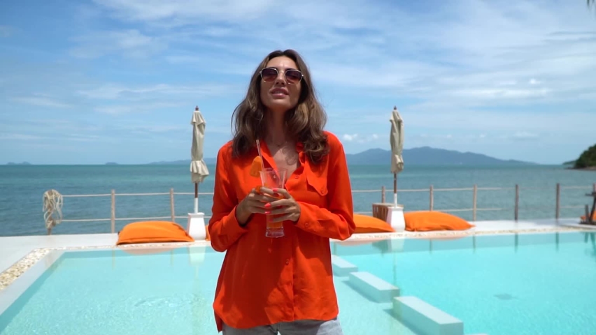 Beautiful woman in orange shirt and jeans holding fresh carrot juice by swimming pool on vacation having fun, smile and laugh. Natural day light at luxury hotel. | Shutterstock HD Video #1094961639