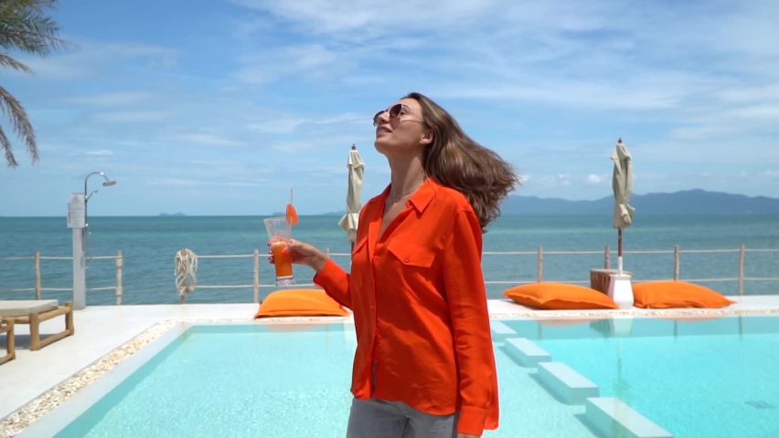 Beautiful woman in orange shirt and jeans holding fresh carrot juice by swimming pool on vacation having fun, smile and laugh. Natural day light at luxury hotel. | Shutterstock HD Video #1094961641
