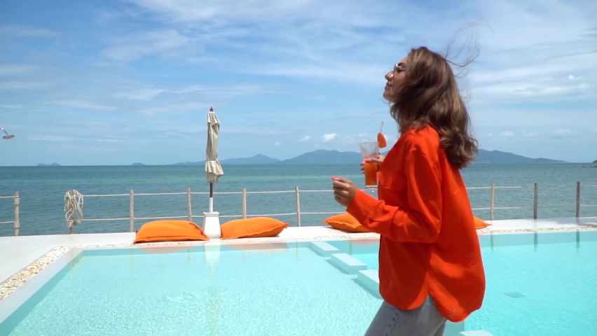 Beautiful woman in orange shirt and jeans holding fresh carrot juice by swimming pool on vacation having fun, smile and laugh. Natural day light at luxury hotel. | Shutterstock HD Video #1094961643