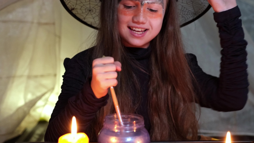 Crazy little witch in black hat is making magic potion and reading spell at night with candle. Kid in costume for Halloween party. Witch stirring soft purple substance in cup and maliciously smiling | Shutterstock HD Video #1094962533