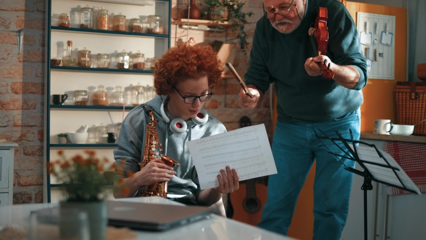 Happy senior man teaching a teenage boy how to play saxophone looking at the notes together. Music student learning saxophone from his old teacher indoor at home Royalty-Free Stock Footage #1094963177