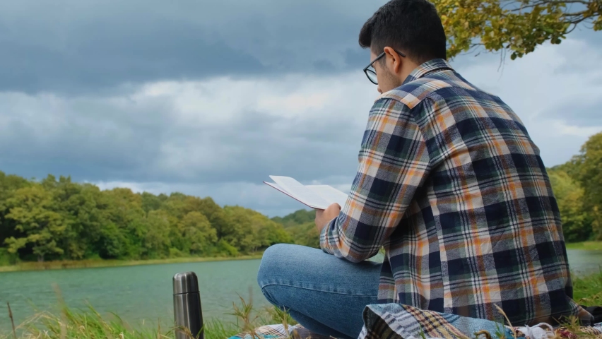 Reading book by the lake in nature, 4k 60 fps video, leisure and enjoyment idea, reading a book concept, man with shirt sitting and read a storybook, peaceful and relaxing activity in forest, cloudy