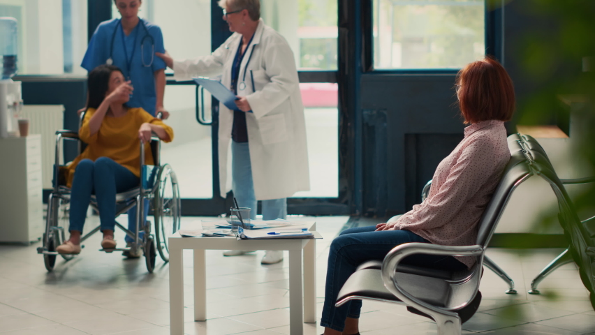 Doctor talking to person with disability and doing checkup visit with asian woman, attending medical appointment in clinic lobby. Receiving support and healthcare advice at consultation. | Shutterstock HD Video #1094968397