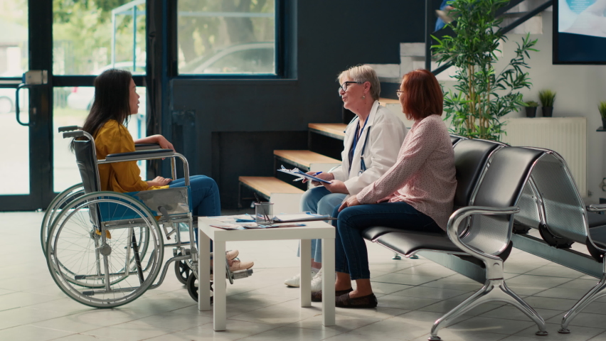 Nurse giving support to patient with impairment in wheelchair, attending medical consultation exam. Asian woman dealing with chronic and physical disability at healthcare clinic. Handheld shot. | Shutterstock HD Video #1094968403