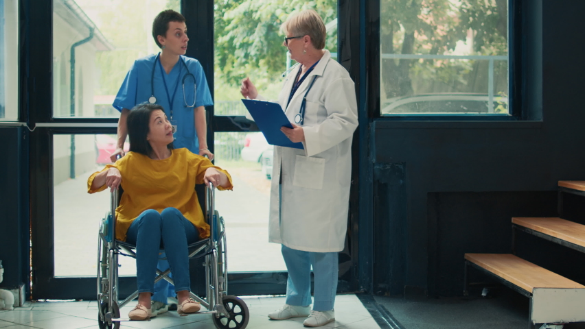 Medical assistant helping patient in wheelchair with impairment, attending appointment at health center. Female wheelchair user suffering from chronic physical disability in lobby. | Shutterstock HD Video #1094968419