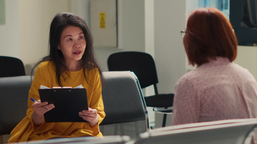 Asian women talking about medical report papers in facility lobby, filling in checkup form before attending exam appointment with physician. Sitting in waiting room at hospital clinic. | Shutterstock HD Video #1094968423