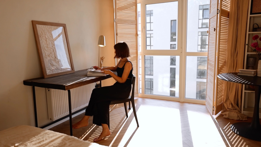 Woman reads some magazin sitting by the table in cozy and sunny living room of modern apartment. Spending leisure time with her dog at home