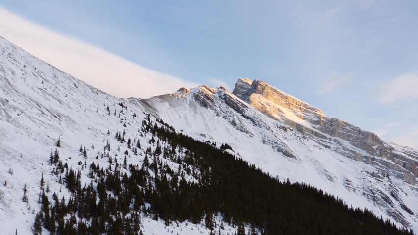 Pan from summit of slanted granite mountain, covered in snow, at sunrise in Canadian Rockies in Canmore Alberta. Royalty-Free Stock Footage #1094975679