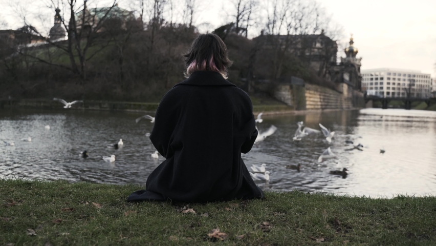 Young woman in a black coat sitting in a park in autumn in afternoon and feeding ducks in a sea | Shutterstock HD Video #1094976213