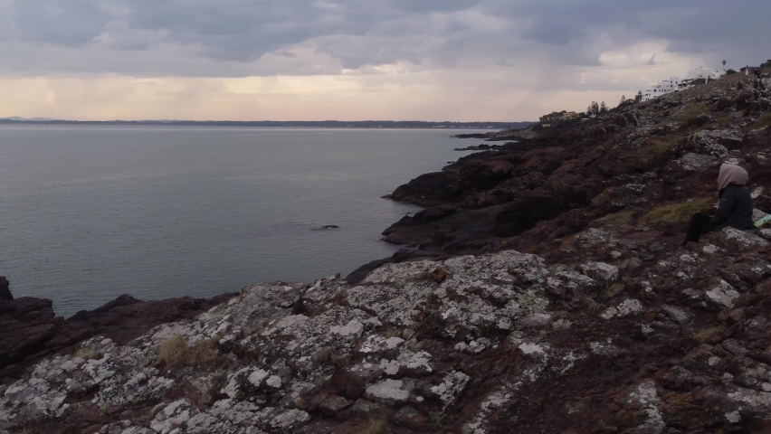 Isolated and unrecognizable woman sitting on rocky cliff looking at seascape panorama, Punta Ballena. Punta del Este in Uruguay. Aerial drone orbiting | Shutterstock HD Video #1094976251