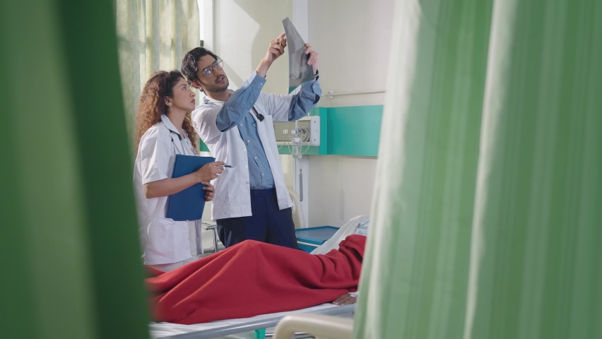 Young Indian Asian Female and Male physician doctors wearing white aprons or lab coat and stethoscope standing discussing the medical record or X-ray report of a patient lying on bed in hospital ward | Shutterstock HD Video #1094977965