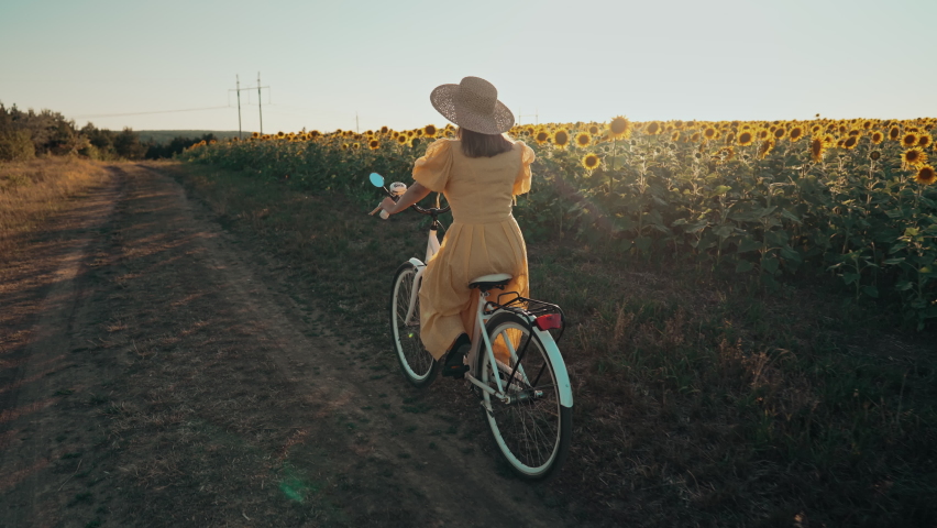 Rural woman in timeless dress riding retro styled white bicycle on country road alone near sunflowers field. Vintage fashion, amazing adventure, countryside activity, healthy lifestyle. | Shutterstock HD Video #1094979803