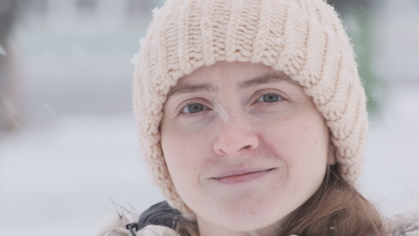 Video portrait of young woman wearing knitted hat outdoors during snowfall. Girl stares intently at camera. Close up.