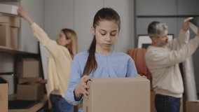 Portrait of the daughter holding the box standing in new flat and her parents lay things out on the background. Family lifestyle and housing concept