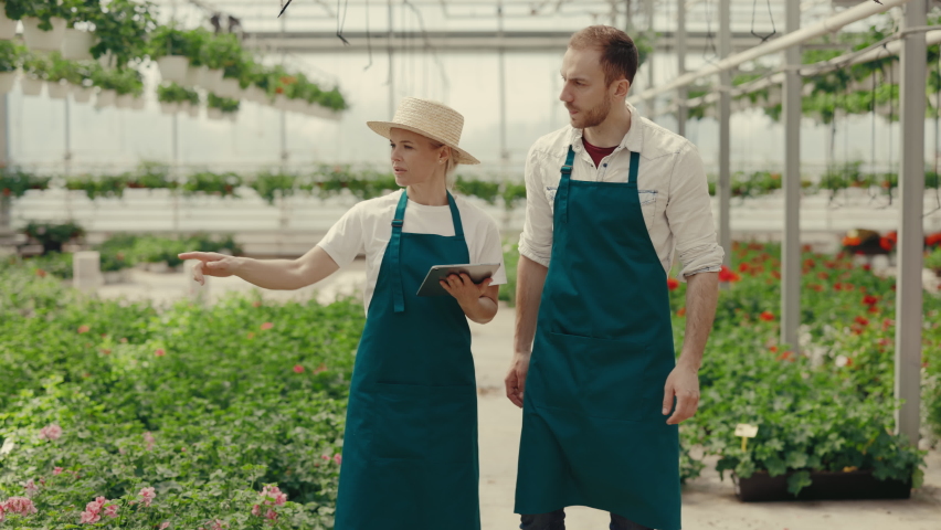 Couple of garden workers are walking through the green house.Female worker showing something to the male worker holding the digital tablet. People and professions concept | Shutterstock HD Video #1094982181