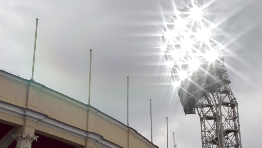 Turning On the Floodlights in the Stadium. Above the open stadium, a large floodlight tower turns on and then off. There are empty flagpoles to place flags | Shutterstock HD Video #1094983597