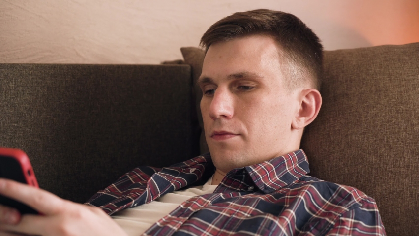 Young brunette man relaxes on brown sofa using gadget, smartphone. internet scrolling. | Shutterstock HD Video #1094984451