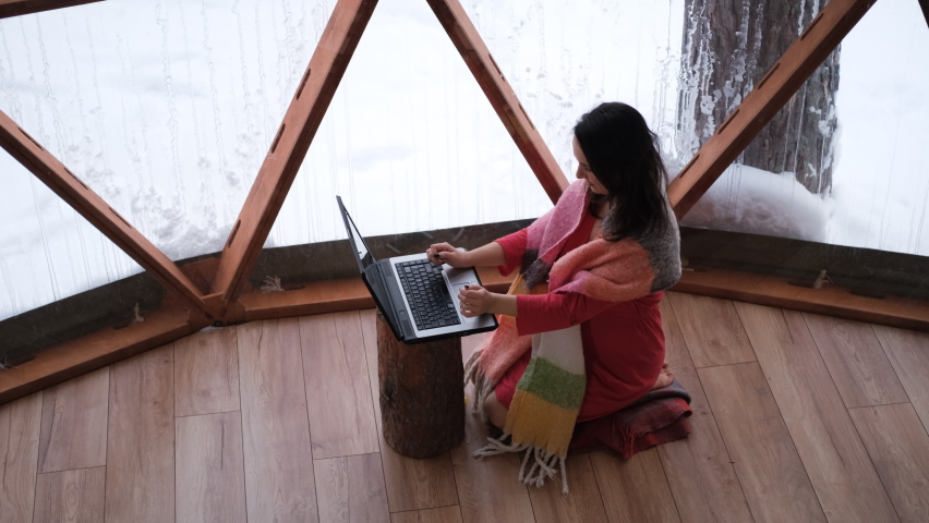Woman working remotely in dome camping in winter, female freelancer using laptop while sitting on the floor of cozy glamping tent over panoramic windows with wild snowy nature  | Shutterstock HD Video #1094984773
