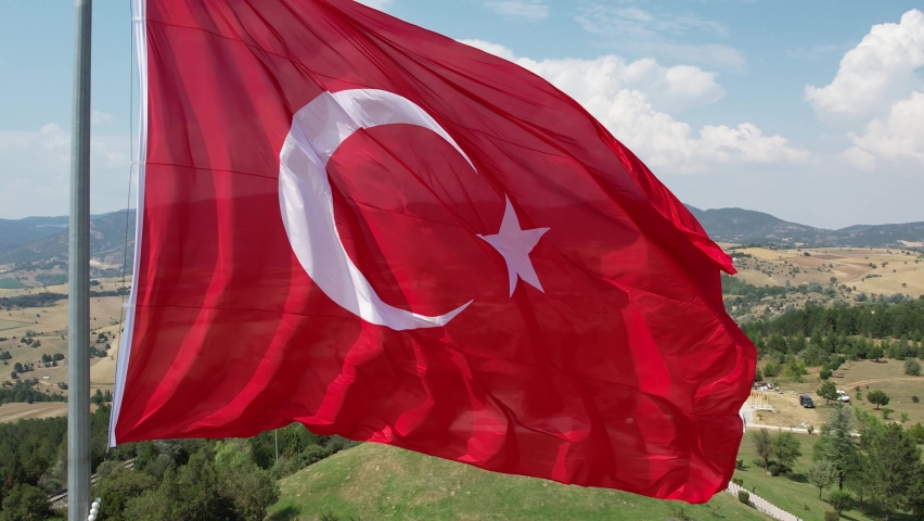 A large bright flag of Turkey flutters on a flagpole high above the ground against the backdrop of the sea, mountains and clouds. Aerial 4k shooting from a drone | Shutterstock HD Video #1094985049