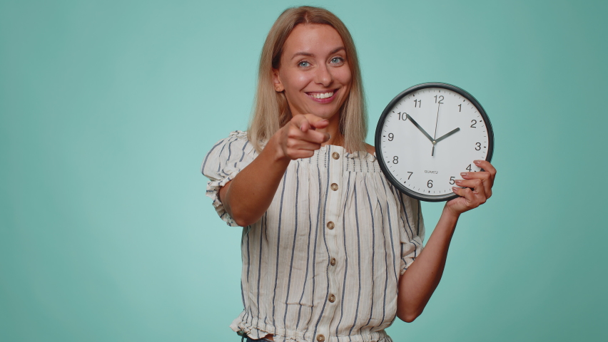 It is your time. Portrait of blonde young woman in shirt showing time on clock watch, ok, thumb up, approve, pointing finger at camera. Adult girl indoors studio shot isolated alone on blue background