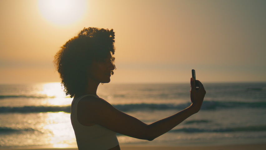 Smiling young woman making selfie at sunrise standing Ursa beach closeup. Cheerful african american girl posing phone camera on beautiful nature. Attractive lady taking photo using smartphone outdoors | Shutterstock HD Video #1094990321