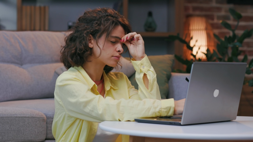 Tired young curly student worker pauses online work, takes off her glasses, massages the bridge of her nose. Burnout, fatigue. overwork at the computer, fatigue, suffering from eye strain | Shutterstock HD Video #1094990793