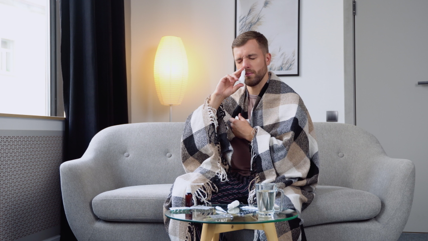 A young man wrapped in a blanket sprays a nasal spray while sitting on sofa