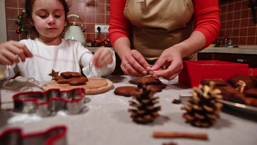 Adorable Caucasian little girl wearing deer antler hoop, holding gingerbread and looking at her mom, who is tying gingerbread cookies with linen rope and stacking them in the gift box. Christmas | Shutterstock HD Video #1094996507