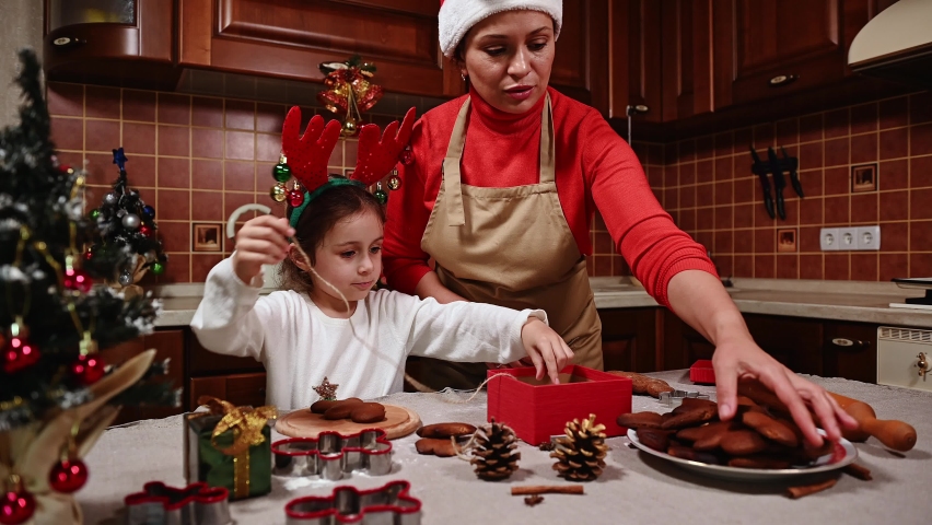 Happy mom and daughter, in Santa hat and deer antler hoop, using linen rope, tying fresh baked gingerbread cookies, putting them in the gift box, enjoying the pastime together in Christmas atmosphere | Shutterstock HD Video #1094996515
