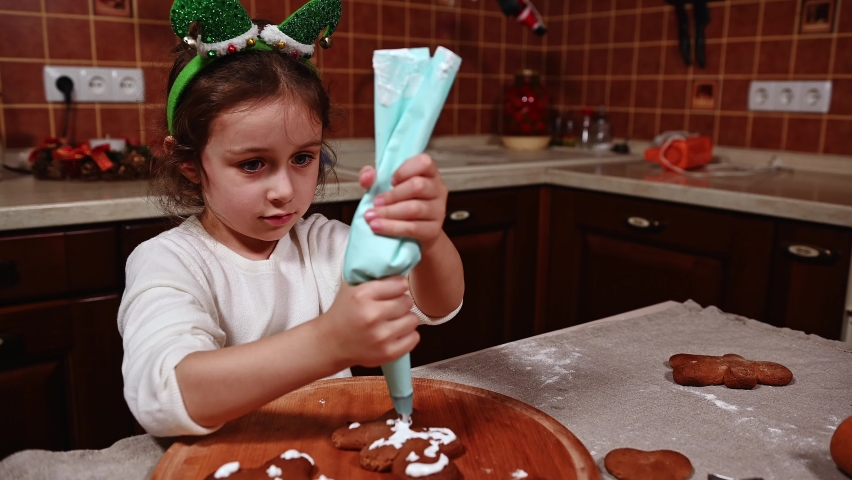 Beautiful child, a lovely Caucasian little girl in white sweater and elf hoop, squeezing icing from a baking bag on baked gingerbread cookies. Child learns decorating pastries with glaze for Christmas | Shutterstock HD Video #1094996517