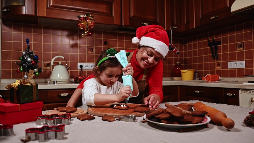 Loving mother wearing Santa hat, standing next to her adorable daughter and teaching her decorating gingerbread cookies for Christmas. Little girl learn cooking and decorating pastries with glaze | Shutterstock HD Video #1094996521