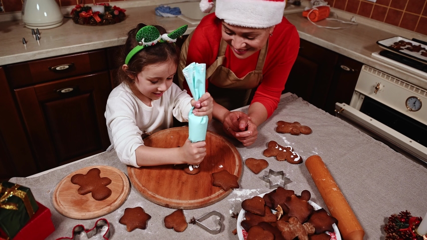 Happy mother in Santa hat, teaches her daughter - a little girl in elven hoop, using icing in baking bag, decorating gingerbread cookies at home kitchen. Christmas. Family. Cooking. Winter holidays | Shutterstock HD Video #1094996527