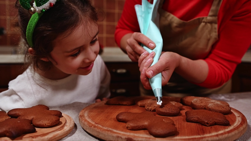 Adorable Caucasian child, little girl in white sweater and an elven hoop, looks at gingerbread cookies while her mom decorates them with icing. Decorating cakes with frosting glaze. Christmas bakery | Shutterstock HD Video #1094996531