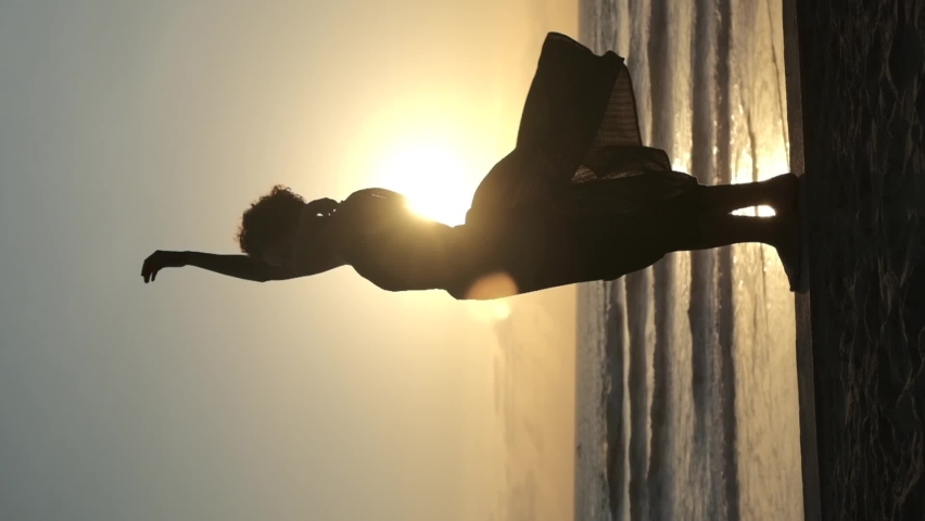 Vertical video of a silhouette of a standing woman in sunlight at dusk. She spins like a little ballerina in a music box. Her dress develops from warm gusts of wind. | Shutterstock HD Video #1095003389