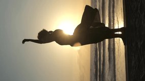 Vertical video of a silhouette of a standing woman in sunlight at dusk. She spins like a little ballerina in a music box. Her dress develops from warm gusts of wind.