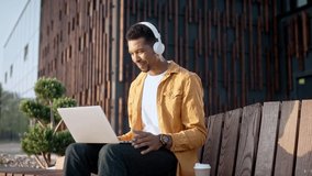 Portrait of African American joyful young man wearing white headphones sitting outdoors and looking at laptop screen having video call online Young male video chatting on street Leisure, communication