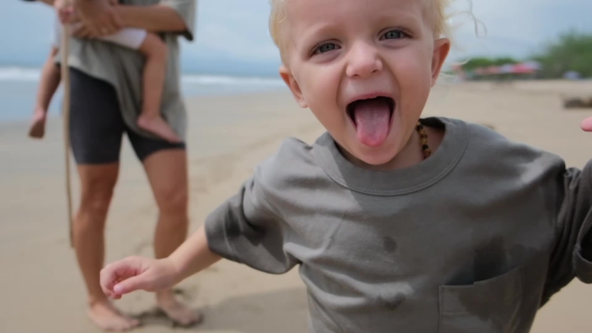 A small blond child runs forward, leaving his mother and sister behind him. The kid laughs and sticks out his tongue while running. He rejoices that he can run fast in his childish imagination. | Shutterstock HD Video #1095005143