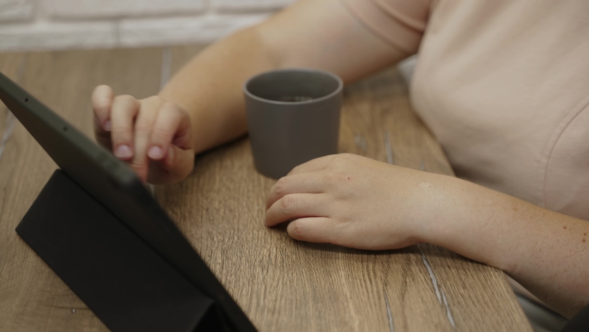 Female hand with natural nail plates without manicure is tapping, swiping on screen of black tablet, close-up. Unrecognizable woman is sitting at the table with cup in the room. | Shutterstock HD Video #1095005163
