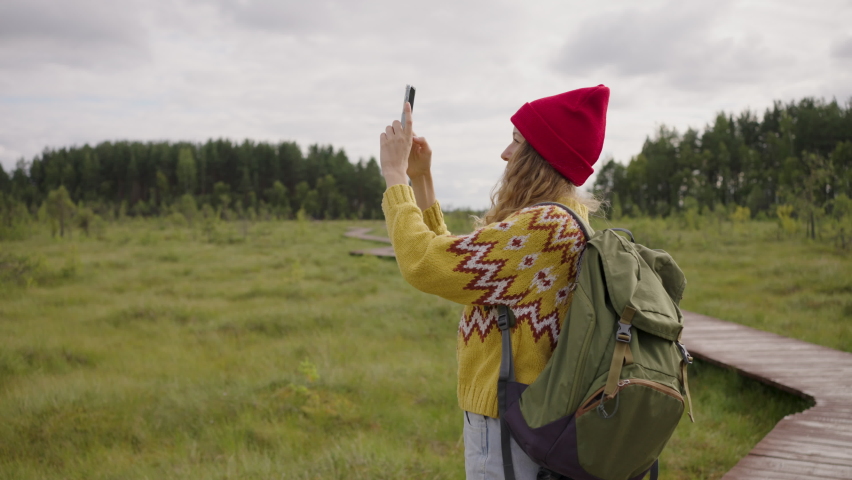 Young woman in red hat takes picture of forest standing on meadow. Hiker with backpack enjoys spending weekend in wild area on cloudy day