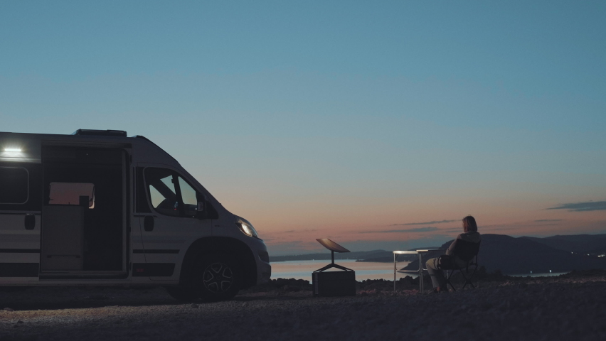 Woman working on her computer and traveling around with her camper van. She gets internet from Starlink SpaceX satellite internet receiver dish. Ocean view at the sunset. Digital nomad, future. Royalty-Free Stock Footage #1095009945