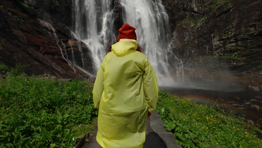 Young girl traveler hiker in a yellow raincoat walks to a waterfall in the highlands