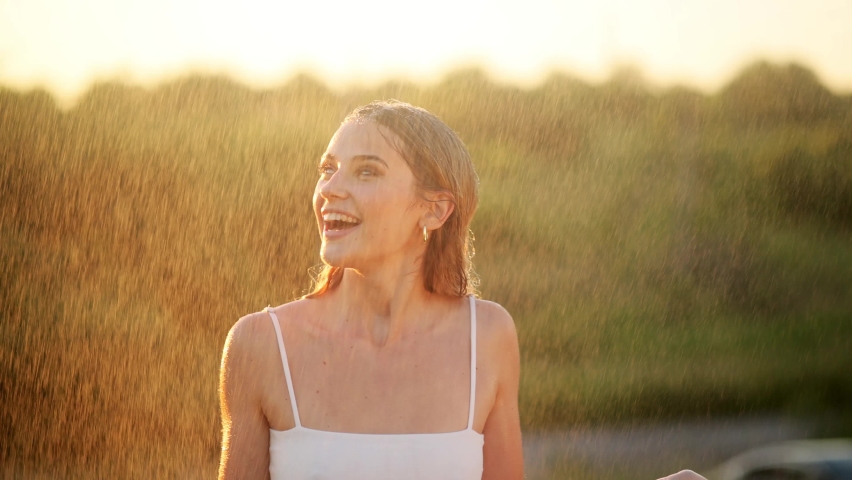 Happy blonde woman having fun in sunny park smiling, fooling around, laughing. Slow Motion. Beautiful caucasian girl dancing under the summer rain. wet hair and clothes. enjoying with drops