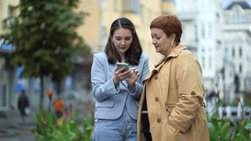 Two women stand on the street of the city and communicate and look at the phone, discuss news, social networks
