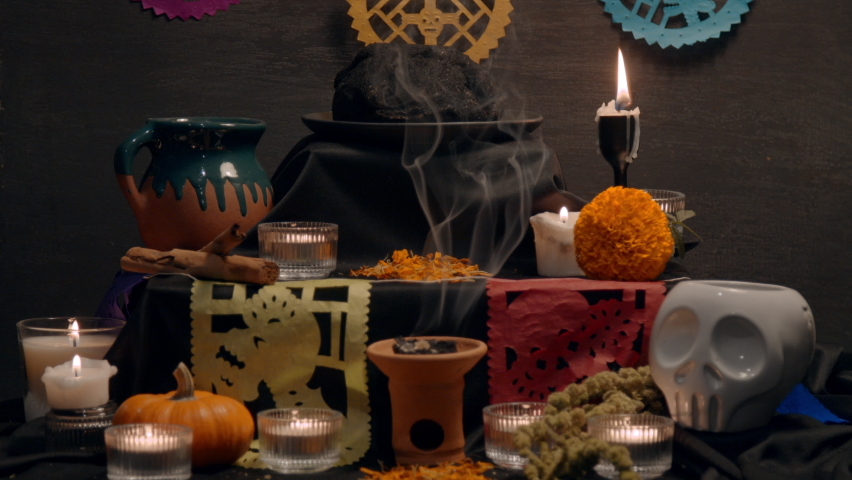 Traditional mexican bread of the dead "pan de muerto" with black ashes in Mexican altar with “papel picado”, copal, incense burner, cempasuchil flowers, coffee, pumpkin, ceramic skull. Slow motion.  | Shutterstock HD Video #1095019543